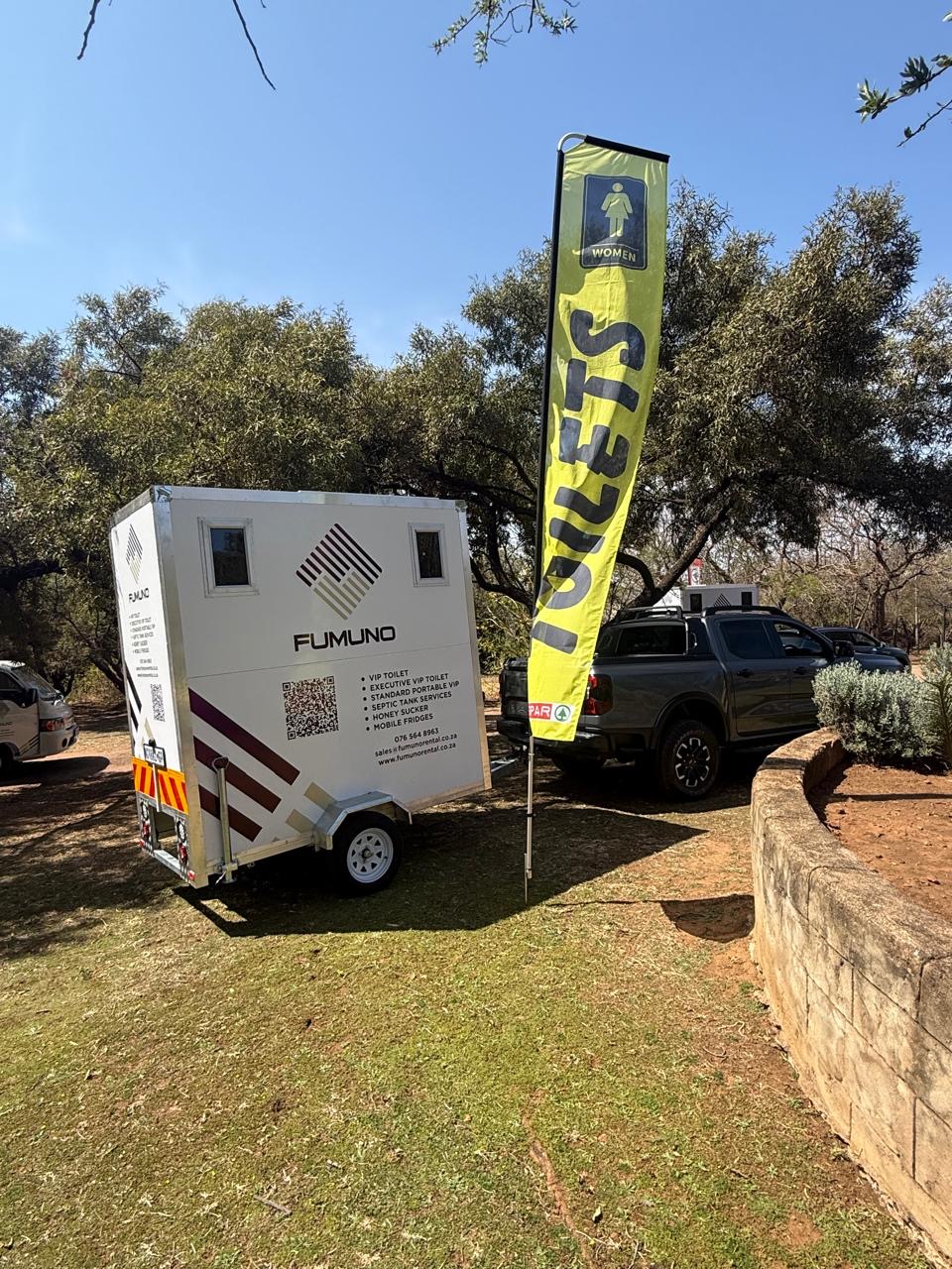 Fumuno branded VIP mobile toilet trailer set up under trees at an outdoor bushveld event in Gauteng, next to a yellow TOILETS feather flag with women's signage.
