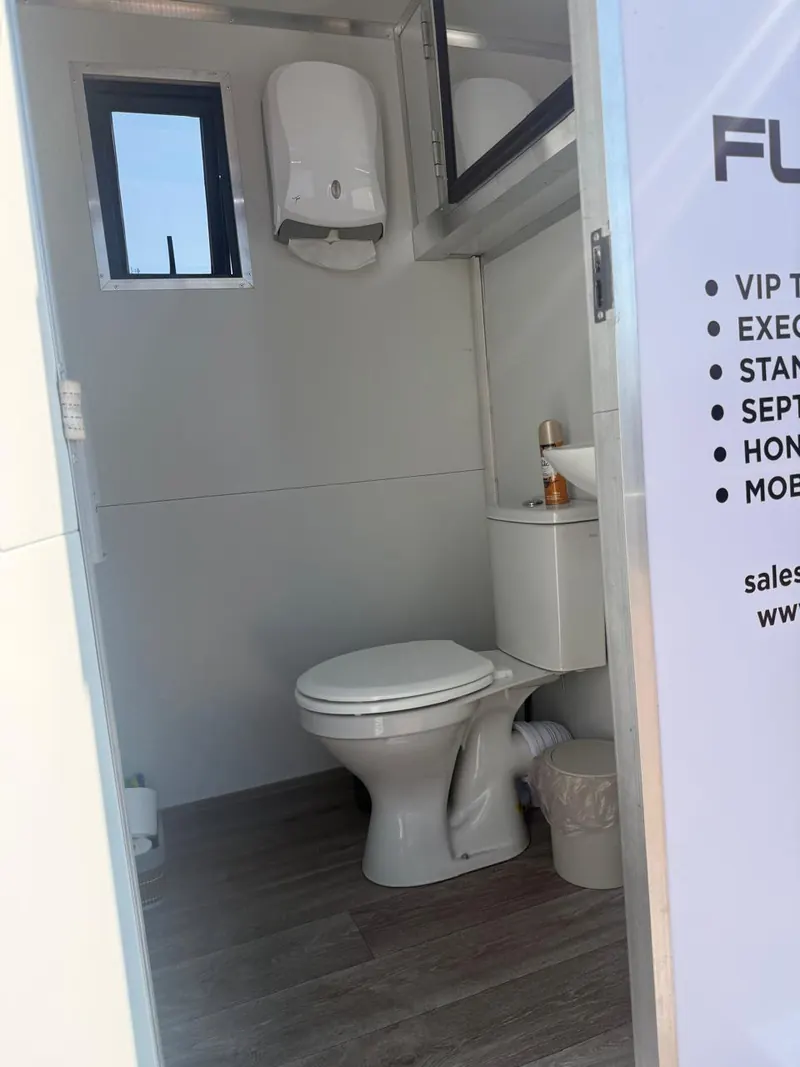 Interior of a Fumuno VIP mobile toilet trailer showing a flushing ceramic toilet, hand basin, paper towel dispenser, mirror, window and vinyl wood-look flooring.