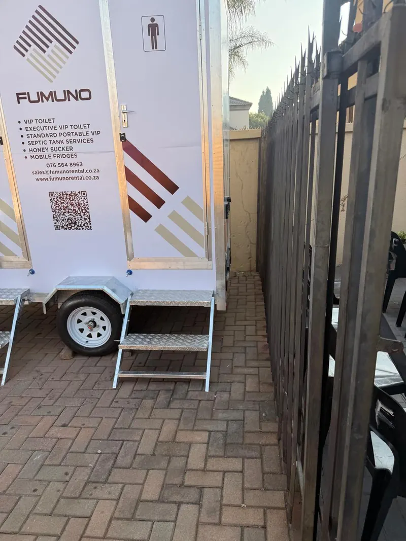 Side view of a Fumuno VIP mobile toilet trailer on a paved driveway showing branded livery, QR code, contact details and galvanised access steps.