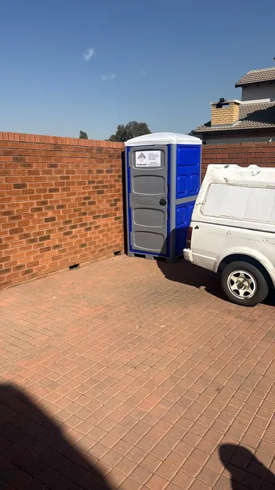 Fumuno blue portable chemical toilet positioned in a brick-walled yard corner next to a delivery vehicle in Gauteng.