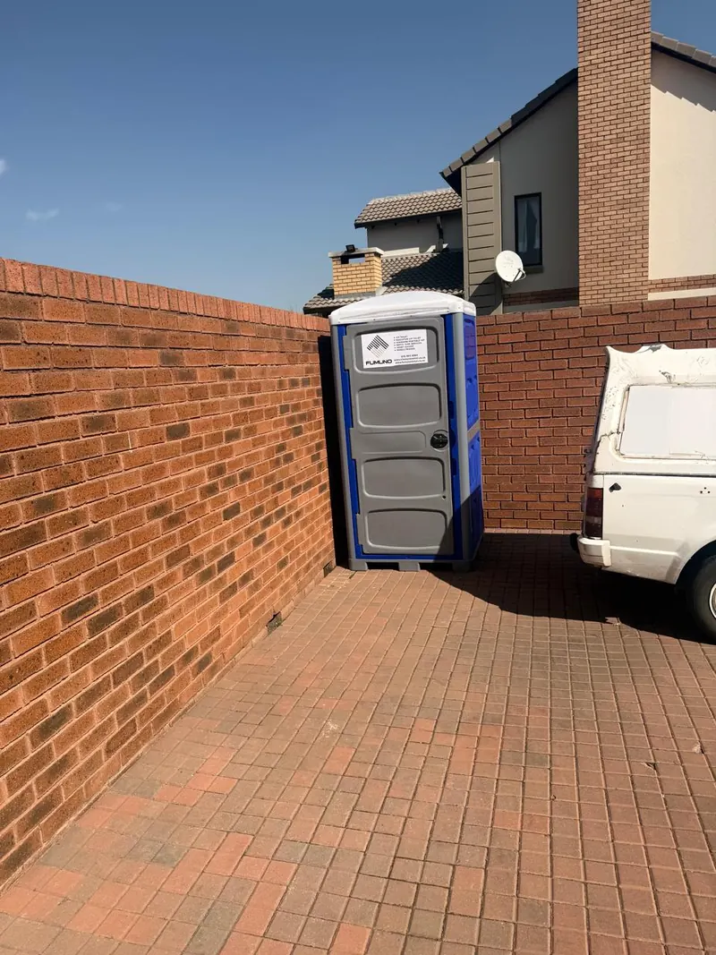 Fumuno portable toilet unit standing beside a face-brick wall and gate on a residential construction site in Gauteng, with the delivery bakkie alongside.