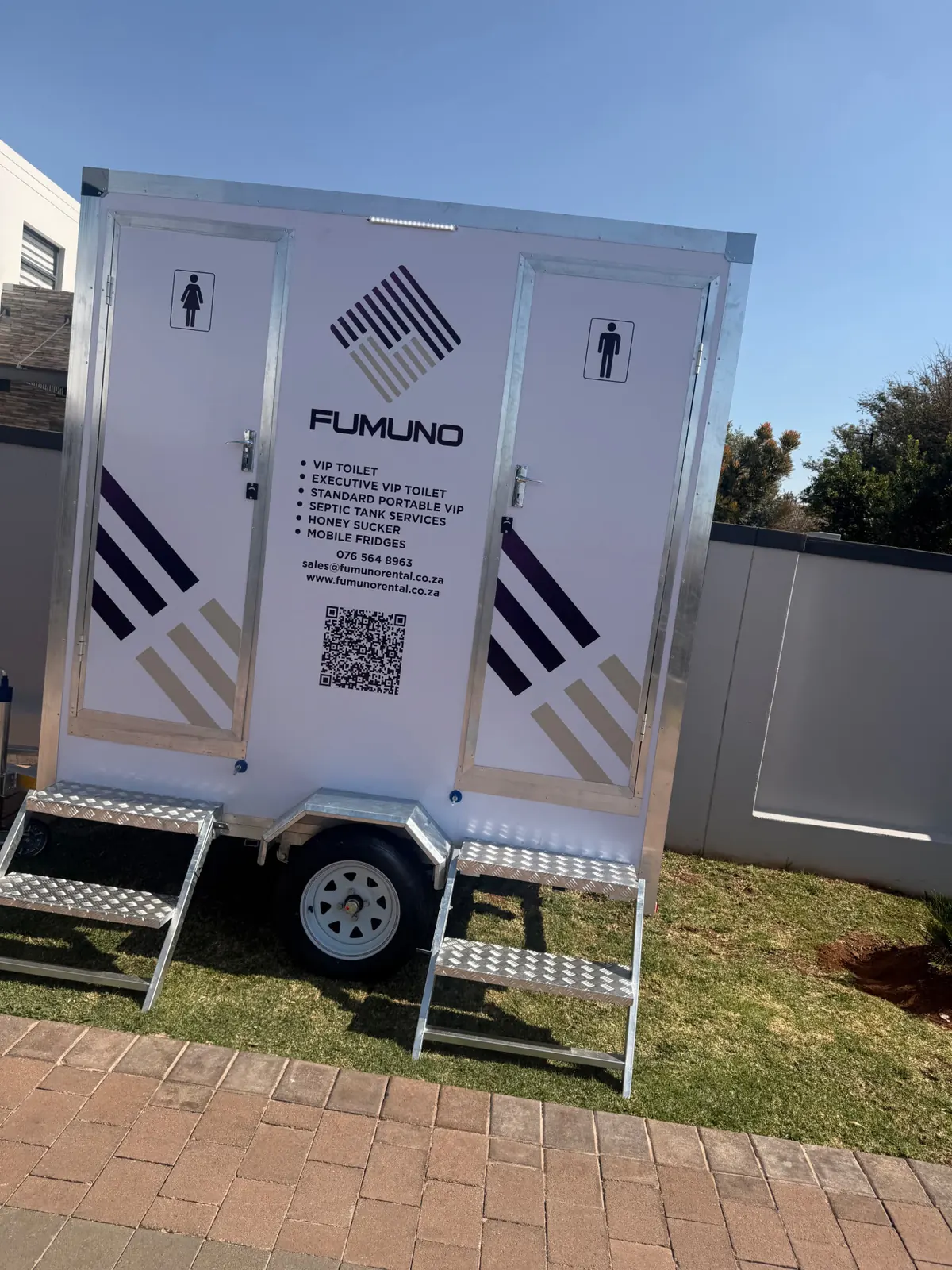 Fumuno twin-door VIP mobile toilet trailer with purple-and-gold branding, male and female door signage and galvanised pull-out steps set up on a garden lawn in Gauteng