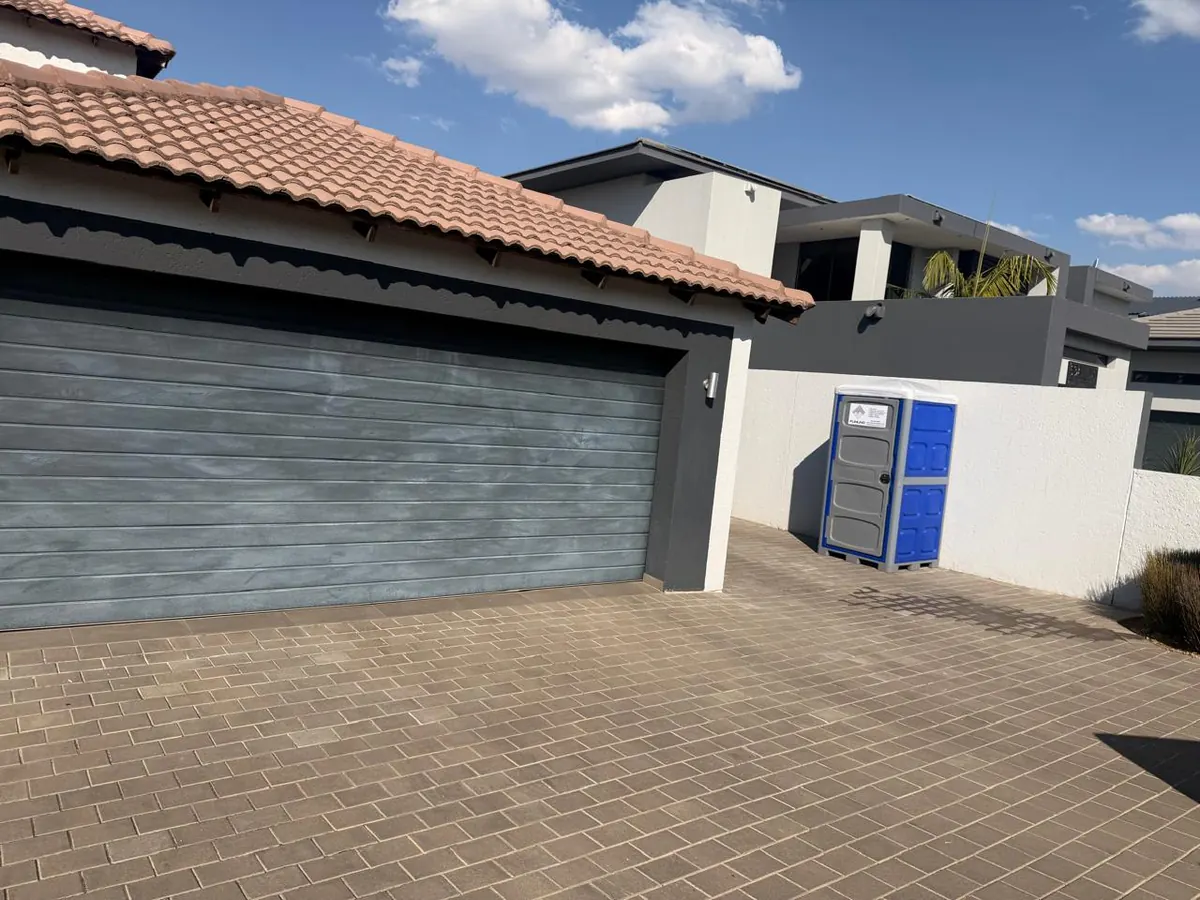 Blue and grey Fumuno-branded standard portable chemical toilet placed neatly against a boundary wall on a paved residential driveway in Gauteng.