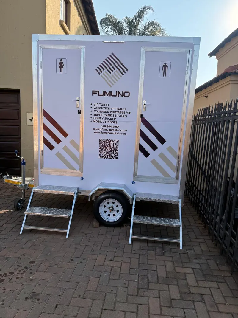 Front view of a Fumuno twin-door VIP mobile toilet trailer with male and female entrances, galvanised steps, branded livery and QR code, set up on a paved driveway in Gauteng.